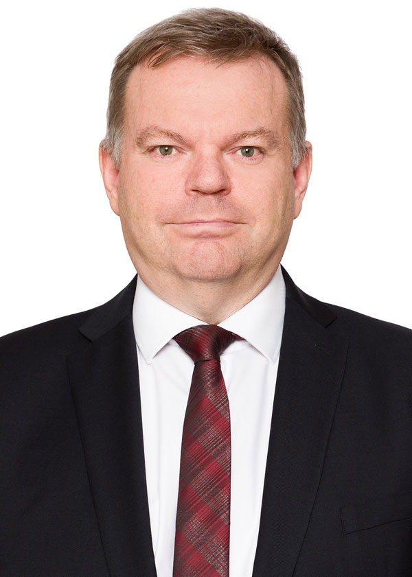 Brian Metson, dressed in a black suit with a white shirt and red plaid tie, stands against a white background. His short hair complements his neutral expression, creating an image of understated elegance.