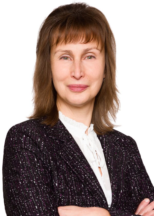 A woman with shoulder-length brown hair, wearing a dark textured blazer and a white blouse, stands with her arms crossed against a white background. She is smiling softly.