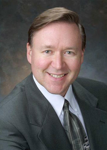 Howard Merricks, dressed in a suit and tie, smiles at the camera against a dark, blurred background.
